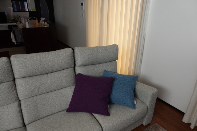 モダンな灰色のソファに紫と青のクッションが置かれ、落ち着いた雰囲気のリビングルーム。ソフトな光を通す黄色のカーテンがあり、リラックスした室内環境を演出している。