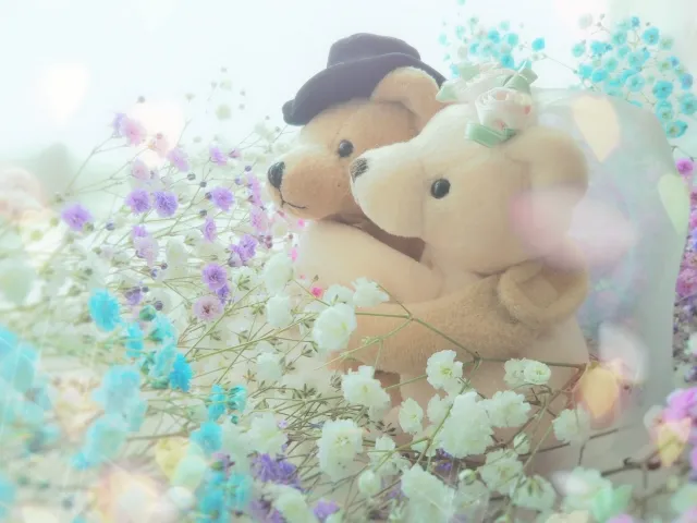 花々に囲まれているクマのぬいぐるみのペアが写されています。一匹は帽子をかぶり、もう一匹は頭にリボンをつけています。柔らかな光の中で、優しい雰囲気を感じさせる写真です。